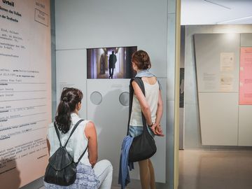 Zwei Frauen, die eine sitzend, die andere stehend, betrachten einen Monitor in der Ausstellung.