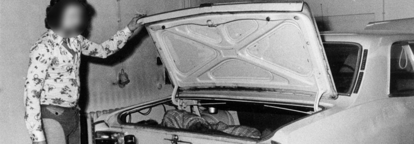 Ein Mann hält den Kofferraum eines Autos auf. Darin befinden sich eine liegende Person.