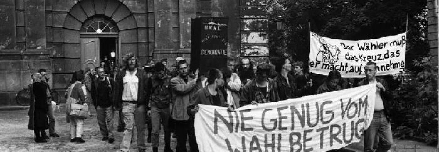 Menschen protestieren mit Transparenten vor einer Kirche gegen den Wahlbetrug bei der Volkskammerwahl 1989.