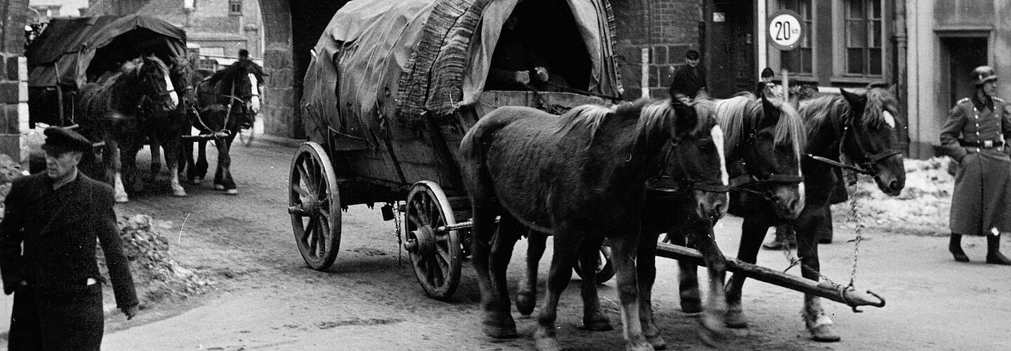 Das Bild zeigt zentral einen von drei nebeneinander gespannten Pferden gezogenen Planwagen, der gerade durch ein aus Backsteinen gebauten Torbogen in eine Stadt hineingefahren ist. Dahinter folgt bereits der nächste Wagen, direkt noch im Torbogen. Während die Wagenlenker nicht zu erkennen sind, gibt es zu beiden Seiten des Wagens und der Straße jeweils einen Mann zu sehen: die Person zur Linken trägt Uniformmütze und langen dunklen Mantel, die Person am rechten Bildrand trägt eine militärische Uniform mit zwei Reihen glänzender Knöpfe und einen Schutzhelm.