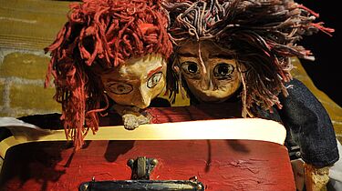 Zwei Handpuppen blicken in einen roten, nicht ganz geöffneten Koffer. Das Innere des Koffers leuchtet leicht.