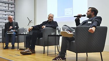 Drei Personen sitzen auf einer Bühne und sprechen miteinander.