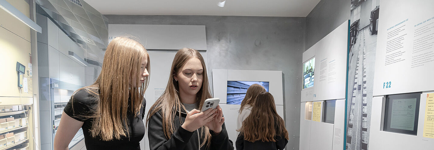 Zwei Personen stehen nebeneinander in einem modernen Museum. Eine Person hält ein Smartphone in der Hand und betrachtet es. Auf der rechten Seite sind Informationswände mit Texten und Bildern zu sehen. Im Hintergrund betrachtet eine Person die Ausstellungsstücke.