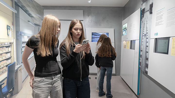 Zwei Personen stehen nebeneinander in einem modernen Museum. Eine Person hält ein Smartphone in der Hand und betrachtet es. Auf der rechten Seite sind Informationswände mit Texten und Bildern zu sehen. Im Hintergrund betrachtet eine Person die Ausstellungsstücke.