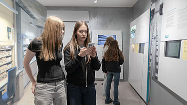 Zwei Personen stehen nebeneinander in einem modernen Museum. Eine Person hält ein Smartphone in der Hand und betrachtet es. Auf der rechten Seite sind Informationswände mit Texten und Bildern zu sehen. Im Hintergrund betrachtet eine Person die Ausstellungsstücke.