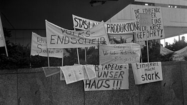 Auf einer Mauer sind mehrere Transparente aufgestellt. Die Losungen, soweit lesbar, lauten: 'Volksentscheid', 'Stasi in die Produktion', 'Ich will kein Soldat sein', 'Gelogen?!! Wir fordern Rechenschaft! Betrogen ?!!', 'Einigkeit macht stark' und 'Freie Wahlen'.