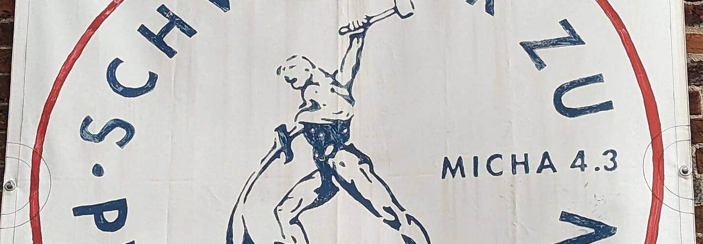 Eine Fahne mit dem Text: Schwerter zu Pflugscharen. In der Mitte des Bildes ein Mann, der aus einem Schwert ein Pflugteil schmiedet