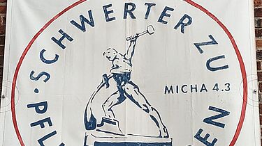 Eine Fahne mit dem Text: Schwerter zu Pflugscharen. In der Mitte des Bildes ein Mann, der aus einem Schwert ein Pflugteil schmiedet