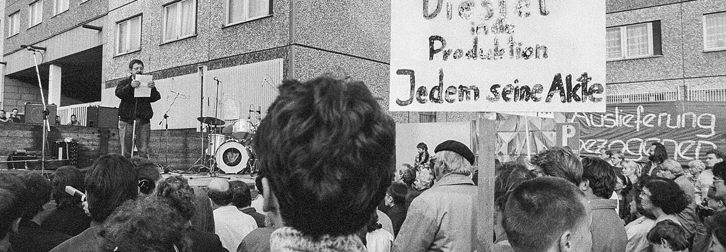 Wolf Biermann spricht vor Demonstrierenden vor der Stasi-Zentrale. Diese halten ein Schild hoch, auf dem geschrieben steht: 'Diestel in die Produktion / Jedem seine Akte'.