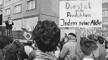 Wolf Biermann spricht vor Demonstrierenden vor der Stasi-Zentrale. Diese halten ein Schild hoch, auf dem geschrieben steht: 'Diestel in die Produktion / Jedem seine Akte'.