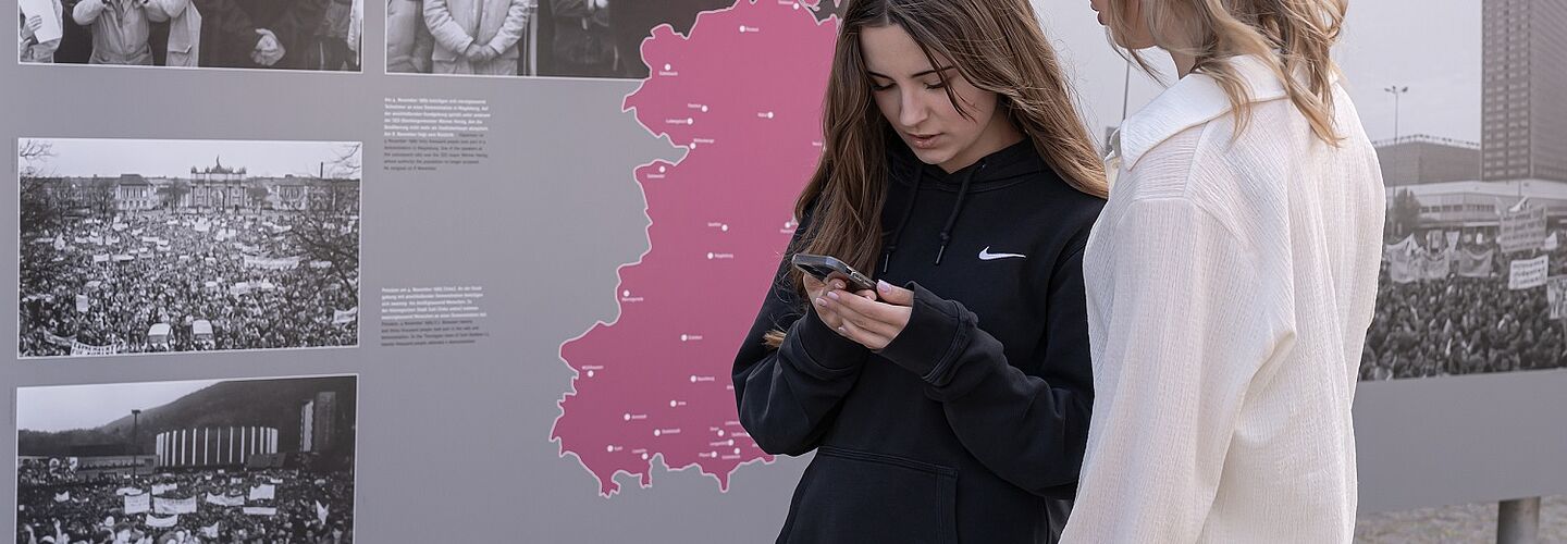 Zwei Schülerinnen stehen in einer Open Air Ausstellung und schauen auf ihre Handys.