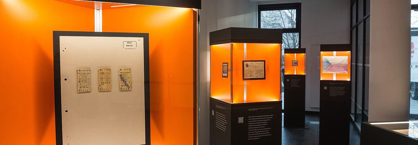 Blick in die Wanderausstellung. Zu sehen sind mehrere Stelen mit Glasvitrinen, in denen sich Dokumente, Fotos und Objekte befinden.