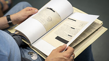 Ein Mann hat auf seinem Schoß eine Akte aufgeschlagen. Es lassen sich Schreibmaschinenschrift und ein Briefumschlag erkennen.