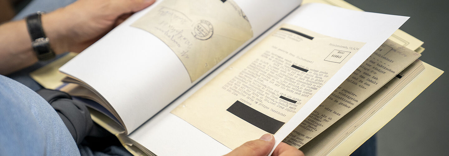 Ein Mann hat auf seinem Schoß eine Akte aufgeschlagen. Es lassen sich Schreibmaschinenschrift und ein Briefumschlag erkennen.