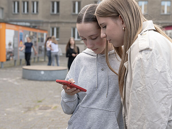 250429_TagderPressefreiheit_MDS_Witzel_3 Zwei Jugendliche schauen auf ein Smartphone, im Hintergrund ist ein Gebäude zu sehen, davor stehen vereinzelt Menschen