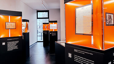 In einem schmalen Raum mit weißen Wänden und schwarzem Boden stehen säulenartig Vitrinen mit schwarzen Sockeln und beleuchteten Glaskästen. Hinter Glas mit orangenem Hintergrung sieht man historische Objekte und Unterlagen aus dem Stasi-Unterlagen-Archiv.