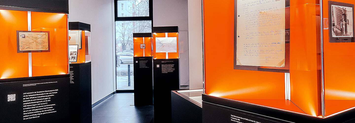 In einem schmalen Raum mit weißen Wänden und schwarzem Boden stehen säulenartig Vitrinen mit schwarzen Sockeln und beleuchteten Glaskästen. Hinter Glas mit orangenem Hintergrung sieht man historische Objekte und Unterlagen aus dem Stasi-Unterlagen-Archiv.