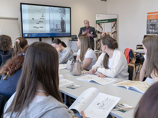 250429_TagderPressefreiheit_MDS_Witzel_1 Eine Gruppe von Schülerinnen sitzt an einem Tisch und verfolgt eine Präsentation auf einem großen Bildschirm. Materialien und Dokumente sind auf dem Tisch verteilt. Eine Flipchart steht neben dem Bildschirm.