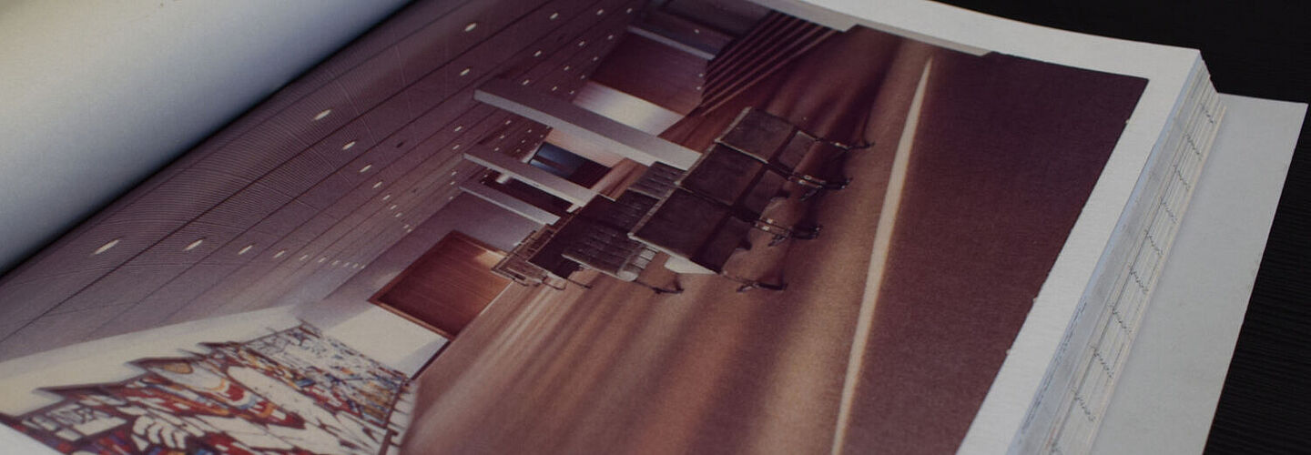 Ein Blick in die aufgeschlagene Akte. Zu sehen ist eine Innenaufnahme eines Saals in Haus 18. Rechts sind Sitzgruppen zu sehen, am linken Rand ein großes Glasmosaik.