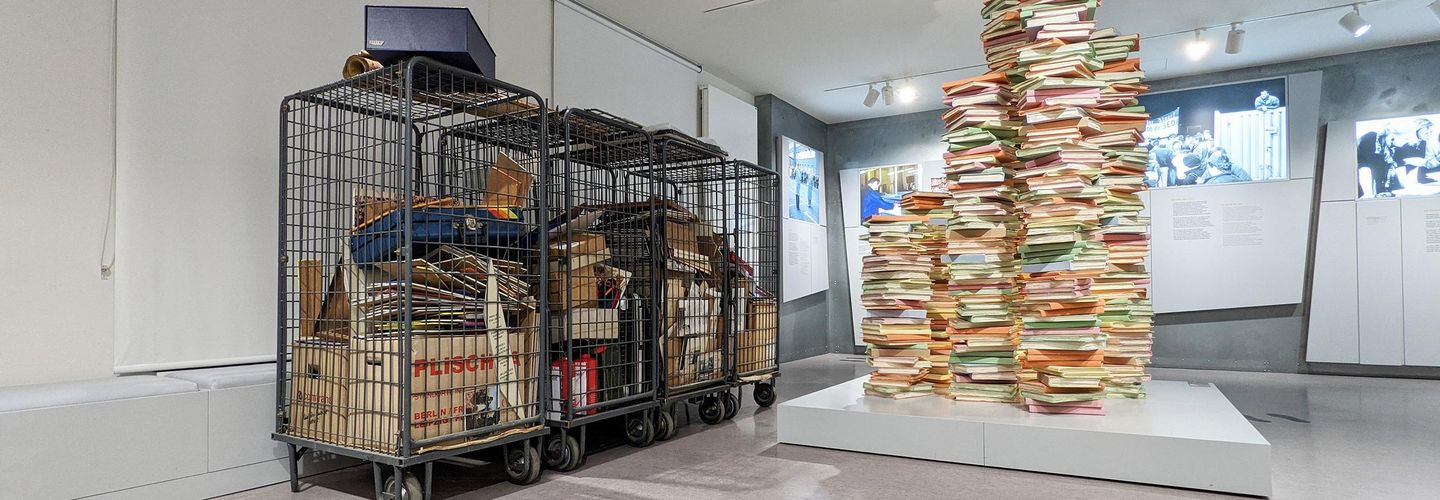 Zwischengelagerte Teile des Traditionskabinetts Auf dem Bild sieht man vier Gitterboxen. In ihnen befinden sich Kartons, Koffer und viele Tafeln. Daneben steht die Skulptur eines Aktenstapels in der Ausstellung.