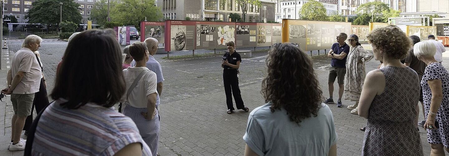 Geländeführung „Einblick ins Geheime“ Eine Gruppe Besucherinnen und Besucher hört einer Vortragenden zu, die eine öffentliche Geländeführung moderiert