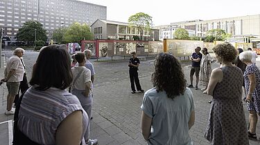 Eine Gruppe Besucherinnen und Besucher hört einer Vortragenden zu, die eine öffentliche Geländeführung moderiert