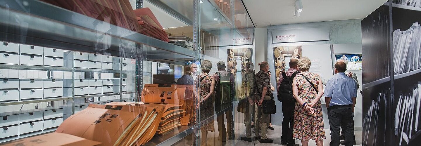 Eine Gruppe von Besuchern in der Ausstellung. Links ist ein Archivregal hinter Glas zu sehen. Die Gruppe spiegelt sich darin.