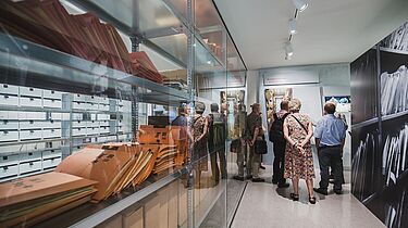 Eine Gruppe von Besuchern in der Ausstellung. Links ist ein Archivregal hinter Glas zu sehen. Die Gruppe spiegelt sich darin.