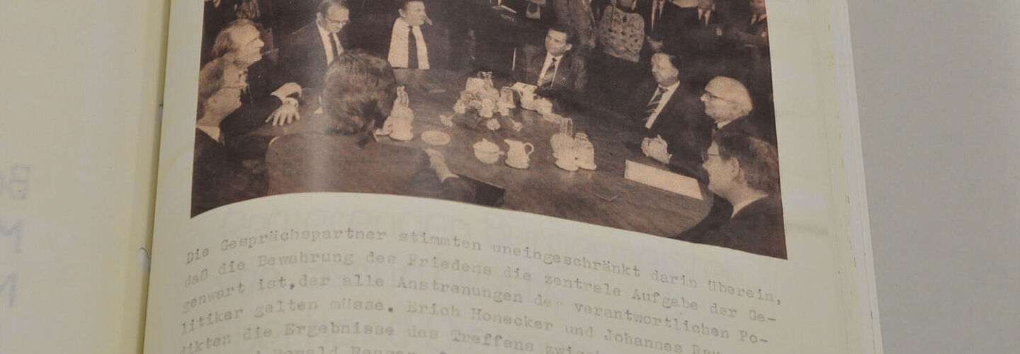 Ein Blick in die aufgeschlagene Akte. Zu sehen ist eine Seite mit einem Foto oben und Text darunter. Das Foto zeigt zehn Männer an einem Tisch und weitere Personen im Hintergrund. Im Text geht es um Gespräche zwischen Johannes Rau und Erich Honecker.