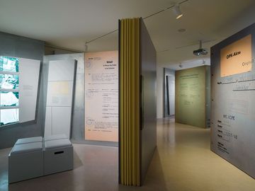 Das Bild zeigt einen Ausstellunsgraum mit mehrere überlebensgroßen Aktenseiten und -deckeln sowie zwei Medienstationen.