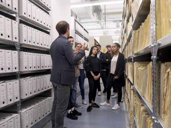 Das Bild zeigt einen Besucherreferenten, der mit einer Gruppe eine Führung in einem Magazinraum im Stasi-Unterlagen-Archiv macht.