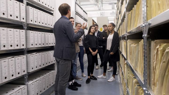 Das Bild zeigt einen Besucherreferenten, der mit einer Gruppe eine Führung in einem Magazinraum im Stasi-Unterlagen-Archiv macht.