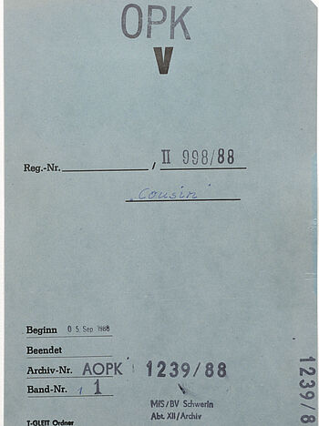Das Bild eines blaugrauen Aktendeckels. Darauf oben zu sehen ist ein Stempel 'OPK', ein großes V, die Registriernummer II 998/88 und das Wort 'Cousin'. Unten zu sehen sind der Beginn des Vorgangs am 05. September 1988 und die Archivsignatur AOPK 1239/88 Band 1. Außerdem der Stempel 'MfS/BV Schwerin, Abt. XII / Archiv.