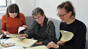 Drei Frauen an einem Tisch. Sie arbeiten mit auf dem Tisch liegenden kopierten Aktenseiten.