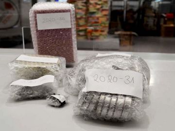 Auf dem Bild sieht man in Luftpolsterfolie verpackte Metallscheiben, so genannte Petschaften. Außerdem sieht man eine verpackte Box. Im Hintergrund ist die Ausstellung zu erkennen.