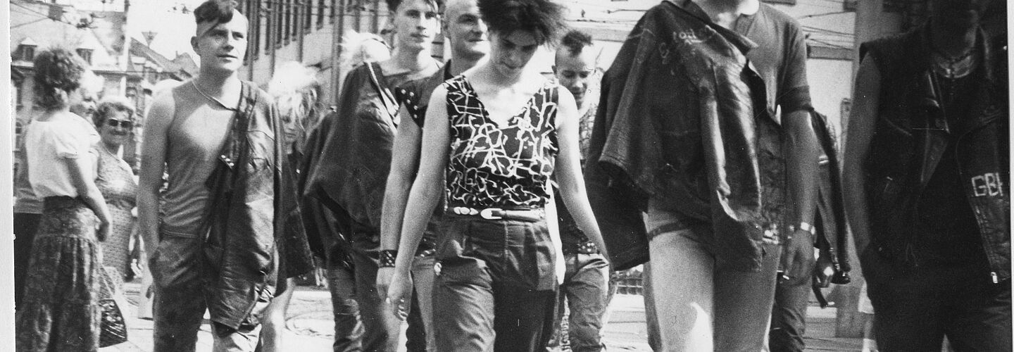 Schwarz-Weiß-Foto mehrerer Personen im Punk-Look auf einer Straße, die an der anscheinend versteckten Kamera vorbei laufen.